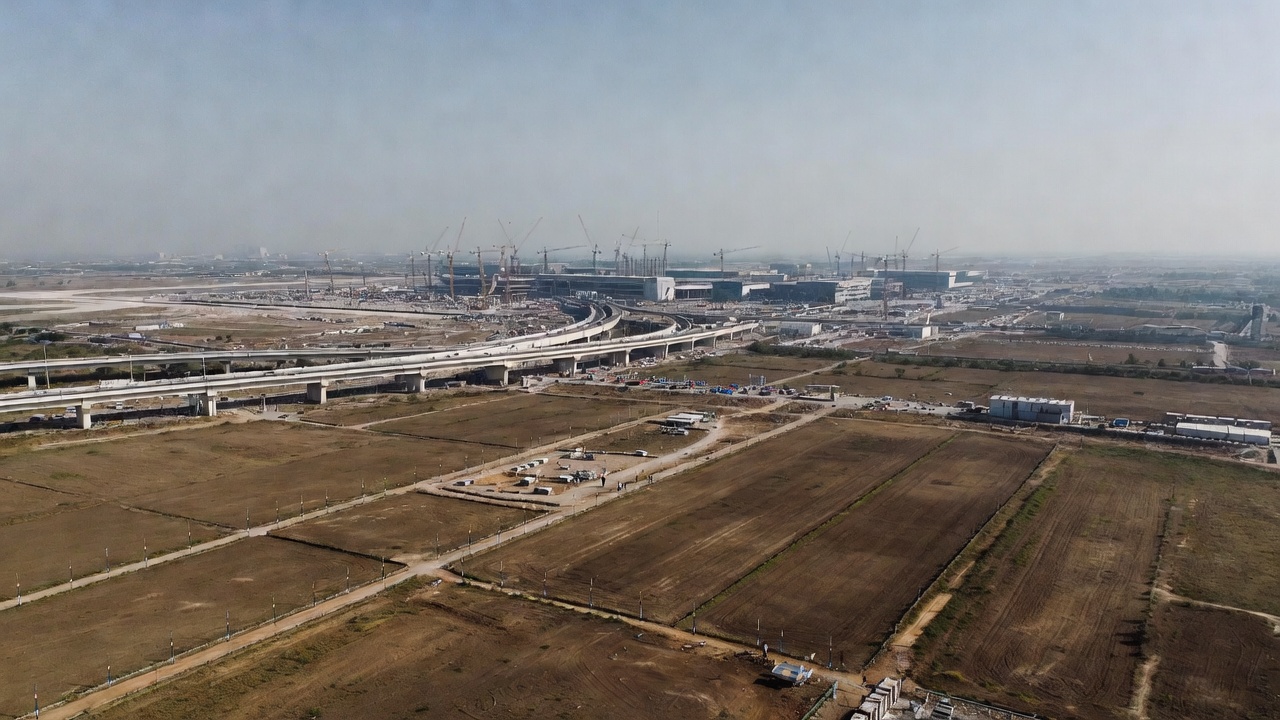 10 Things to Consider Before Buying Land Near Jewar Airport- A wide-angle landscape view of the developing region around Jewar Airport in NCR, India. In the foreground, open land parcels with boundary markers and survey poles. In the background, large airport construction with cranes, runways, and expressway connectivity. Investors and real estate professionals reviewing site maps on a tablet. Bright daylight, realistic photography style, ultra-detailed, 16:9 ratio, cinematic composition, modern infrastructure blending with rural land.
